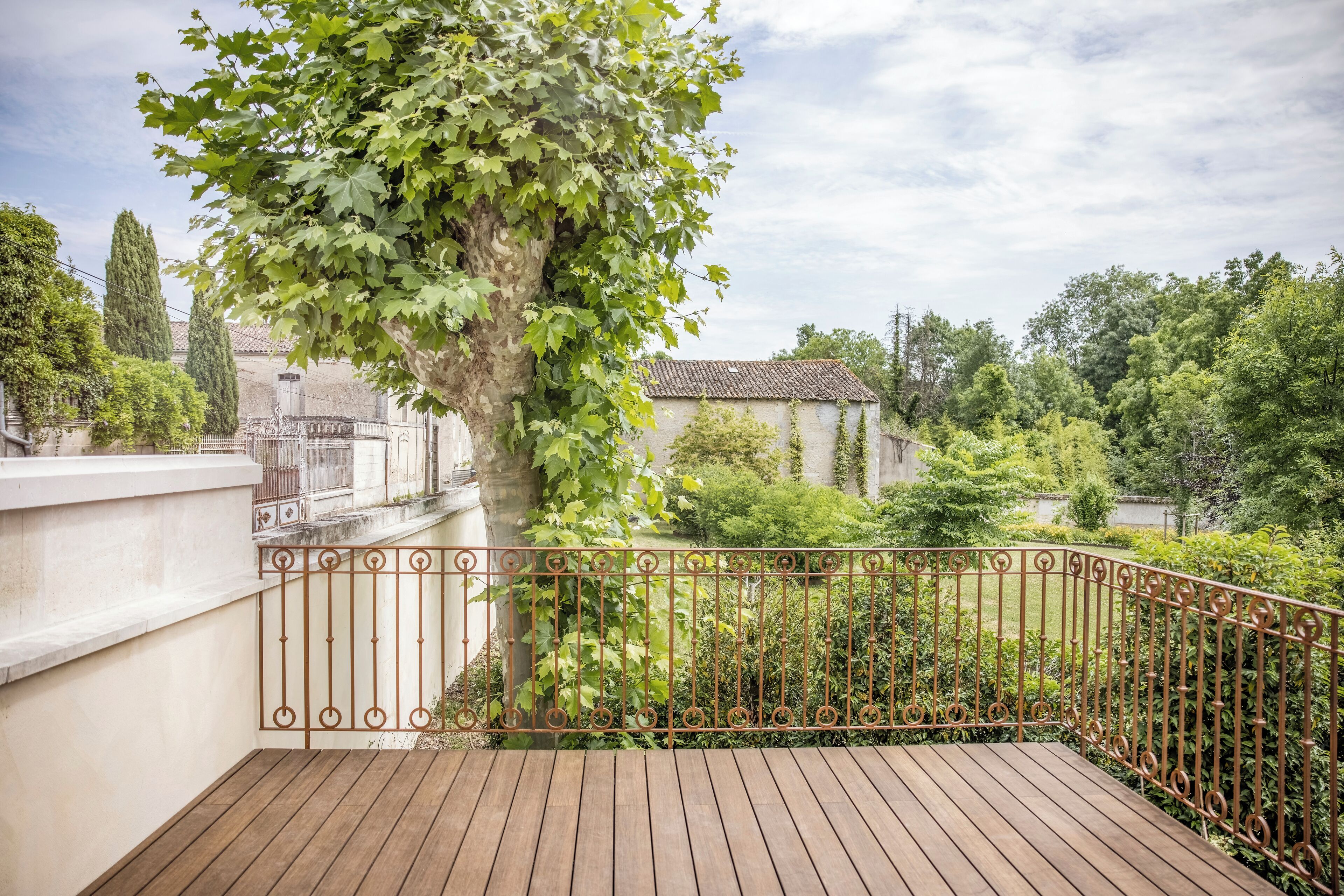 Une terrasse en bois agrémentée d’une balustrade en fer forgé, entourée d’une végétation luxuriante et offrant une vue sur un bâtiment dont le toit est recouvert de tuiles.