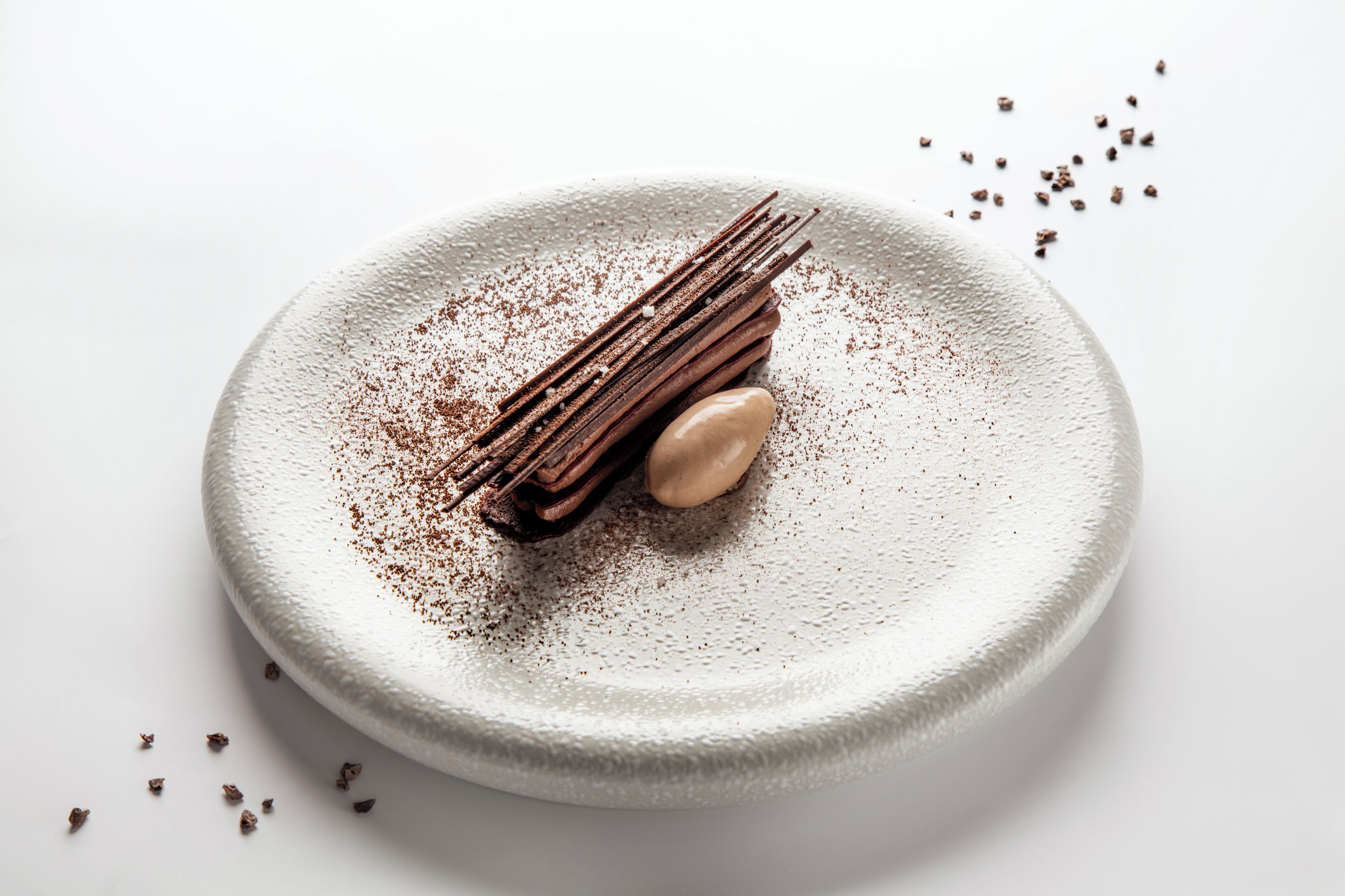 Un dessert servi dans une assiette à surface texturée, accompagné de chocolat et d’une quenelle de crème, le tout agrémenté de morceaux de chocolat éparpillés.