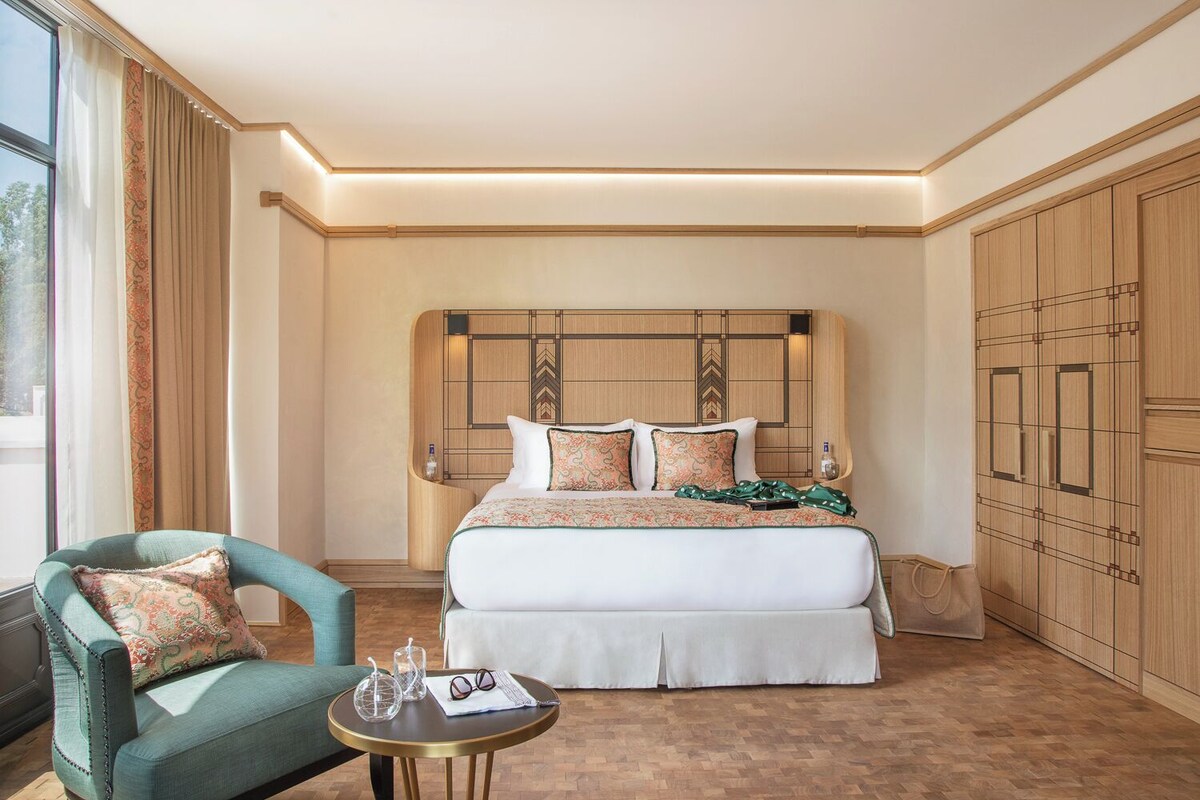 Een hotelkamer met een groot bed, een groene fauteuil, een klein rond tafeltje met glazen en een fles, en een kledingkast.