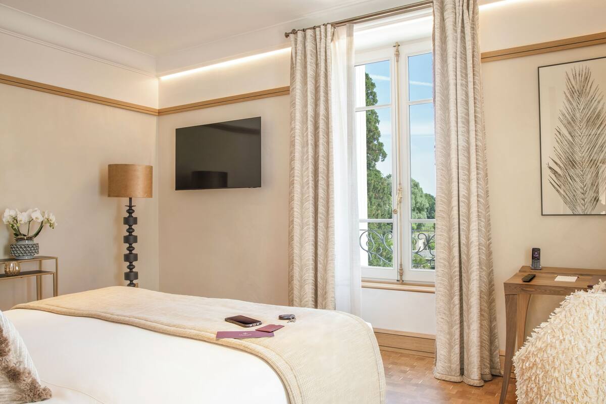Une chambre d’hôtel comprenant un lit, un téléviseur fixé au mur, une fenêtre avec des rideaux, une petite table avec une lampe et une œuvre d’art encadrée accrochée au mur.