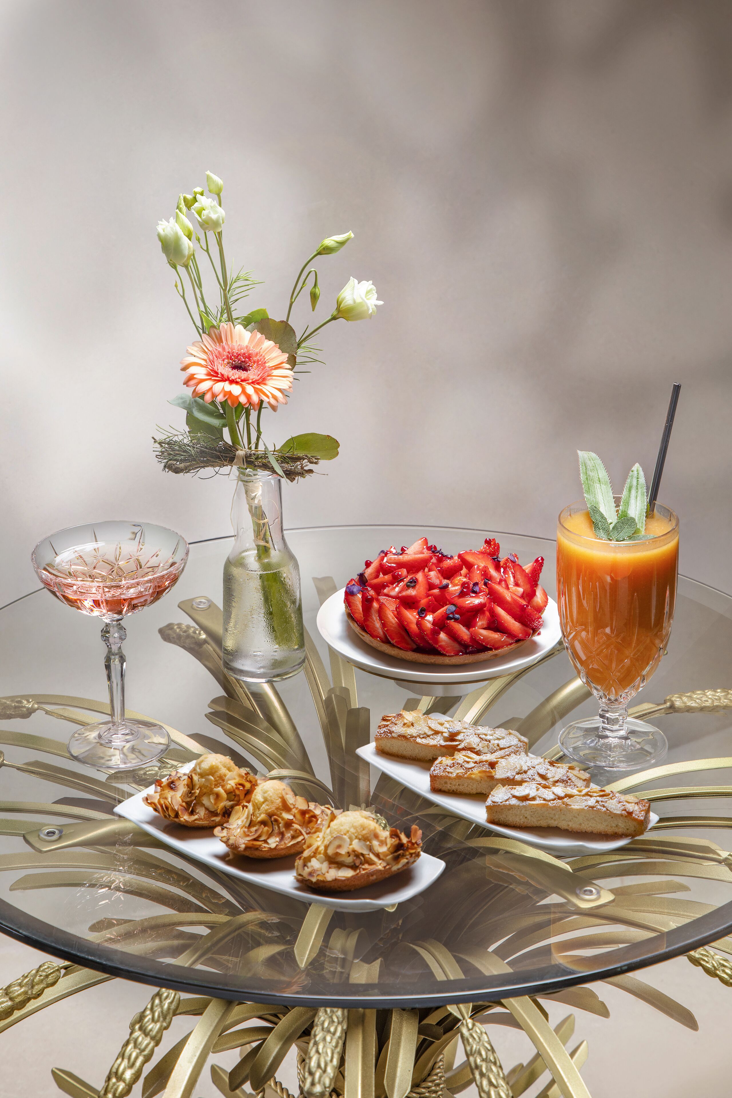 Une table ronde en verre, agrémentée d’un arrangement floral, une assiette de fraises, un verre de jus d’orange et un verre d’une boisson rose.