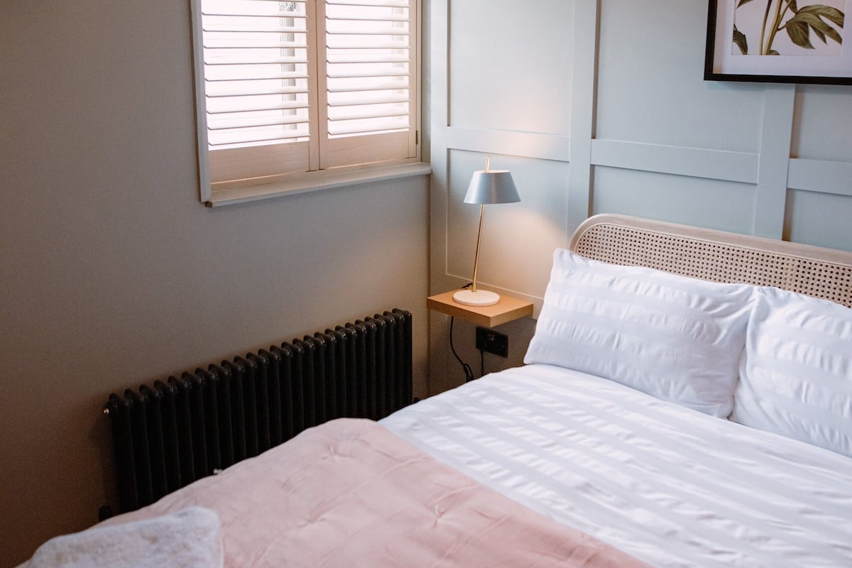 Ein Schlafzimmer mit Bett, Nachttisch mit Lampe, Heizkörper, Fenster mit Jalousien und einem gerahmten Bild an der Wand.