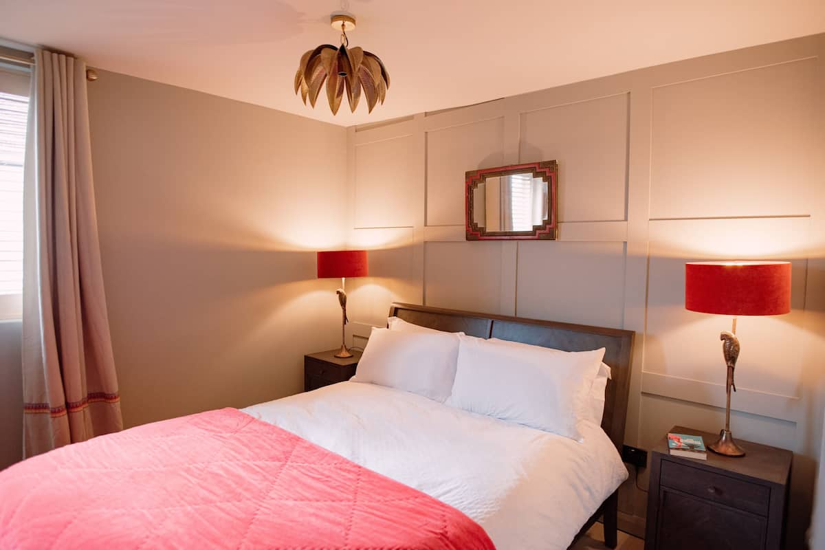 Ein ordentlich bezogenes Bett mit einer rosa-weißen Decke, zwei Nachttische mit roten Lampenschirmen, ein Holznachttisch, ein Spiegel an der Wand und eine Deckenleuchte.