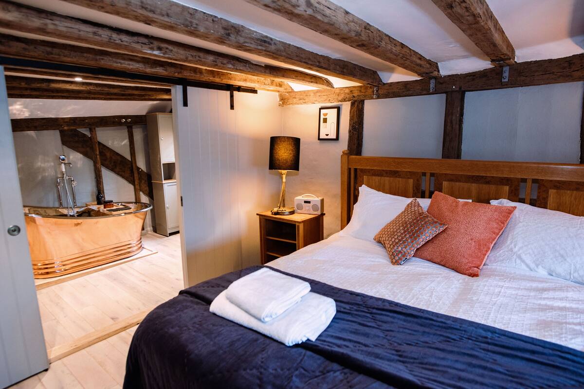 Ein Schlafzimmer mit Holzboden, einem Bett mit weißen und orangefarbenen Kissen, einem Nachttisch mit einer Lampe sowie einer Holzbade Wanne.