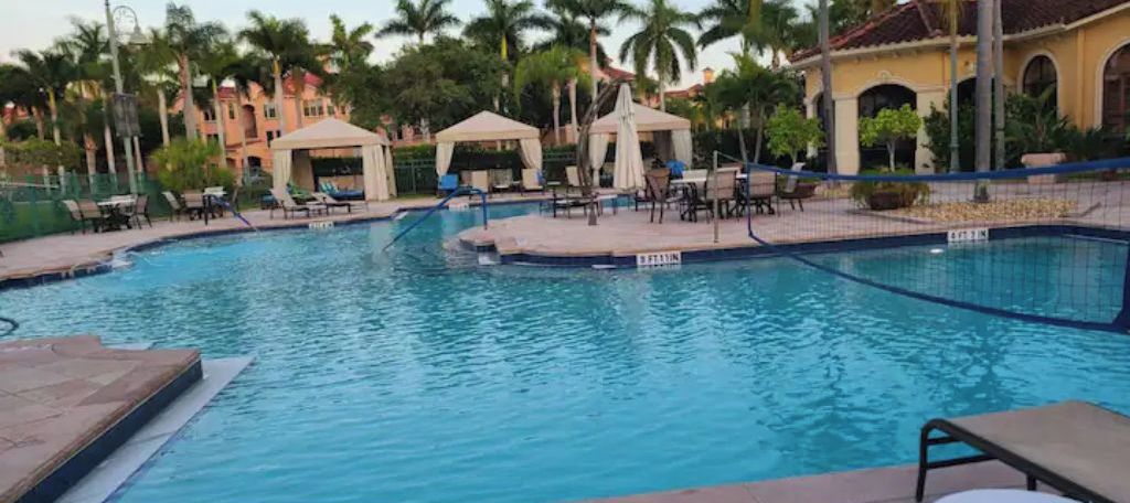 Una piscina con sillas de descanso y sombrillas, rodeada de palmeras y un edificio con arcos.