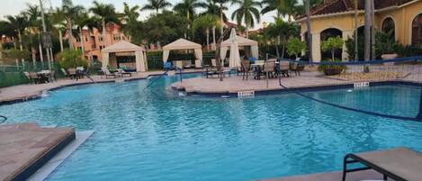 A swimming pool with lounge chairs and umbrellas, surrounded by palm trees and a building with arches.