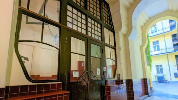 A building entrance with a tiled facade, a dark wooden door, and a view of a yellow building in the background. - Charming 1BR Condo with A/C in Central Budapest - Skyline House (Budapest)