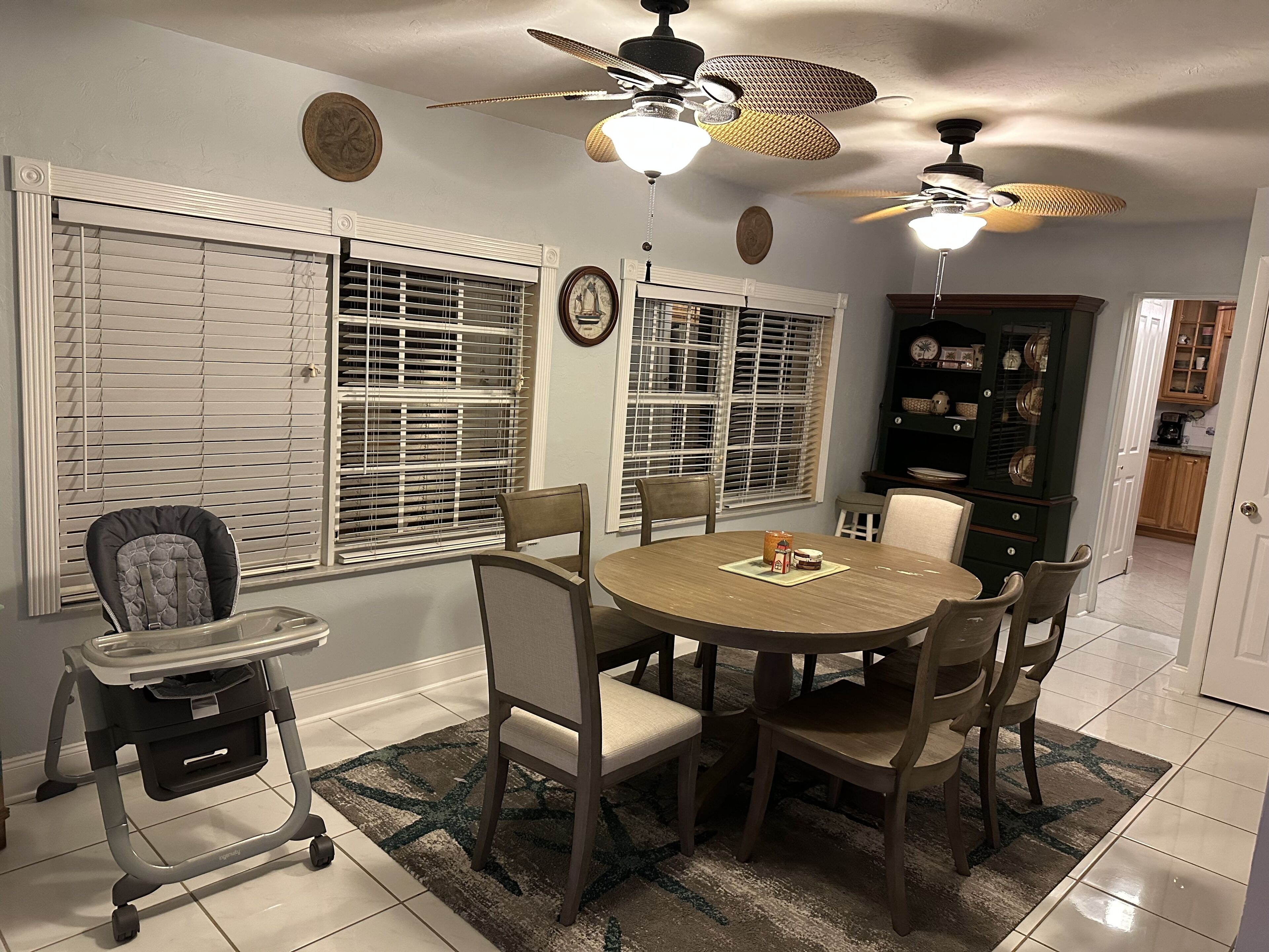 A dining area with a round table, chairs, a ceiling fan, and a kitchen in the background.