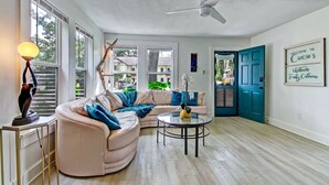 A modern living room with a curved sofa, a round coffee table, and a ceiling fan. There is a door with a sign reading "Welcome to Coco's Uptown Funky Cabana."