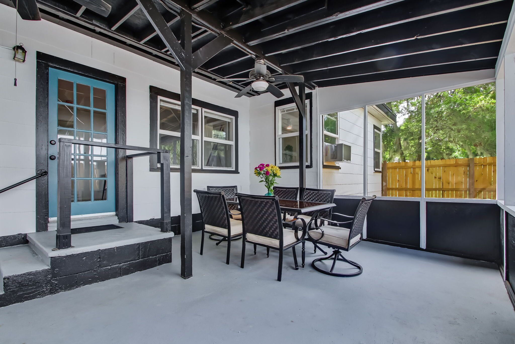 Un patio techado con una mesa y sillas, una puerta azul y una valla de madera al fondo.