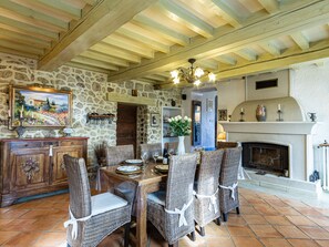 Un coin repas agrémenté d’un mur en pierre, de poutres en bois au plafond, de chaises en osier, d’une cheminée et d’une table dressée pour quatre personnes.