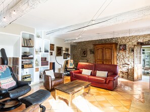 Un vaste salon avec un canapé en cuir rouge, une table basse en bois et un fauteuil en cuir noir. Au fond, on aperçoit un mur en pierre et une armoire en bois.