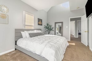 Quarto com cama grande, janela com persianas, lustre, espelho e porta que dá para outro cômodo.