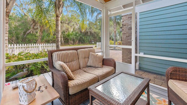 A covered patio with wicker furniture, a glass table, and a view of a white picket fence and greenery. - Island Sun House - Walk to State Park and Beach! (St. Augustine)