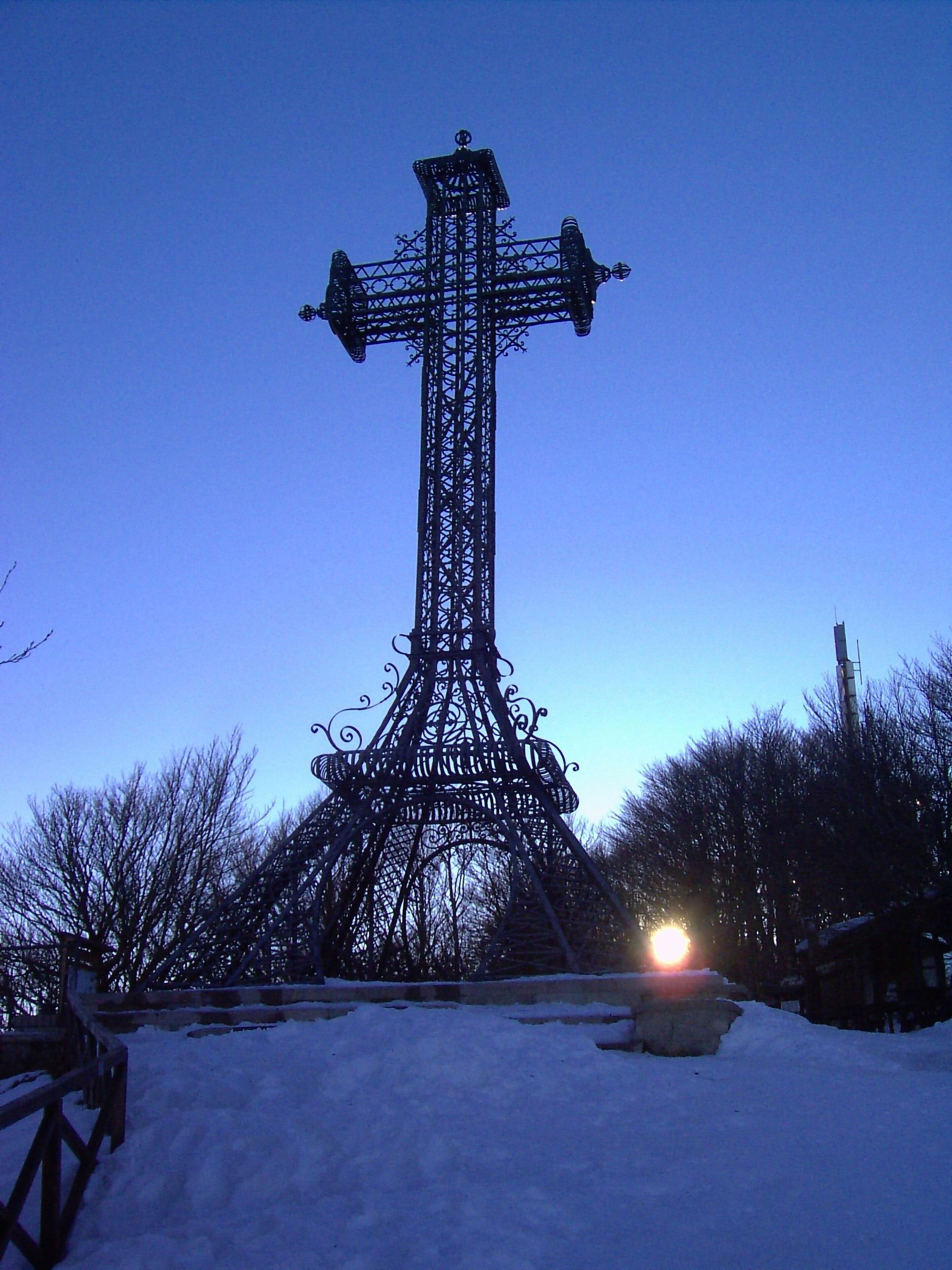 A tall, ornate metal structure resembling a cross, surrounded by snow and trees, with a bright light at its base.