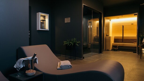 Een moderne spa-ruimte met een ligstoel, een klein tafeltje met een schaal noten en een glas, en een potplant.
