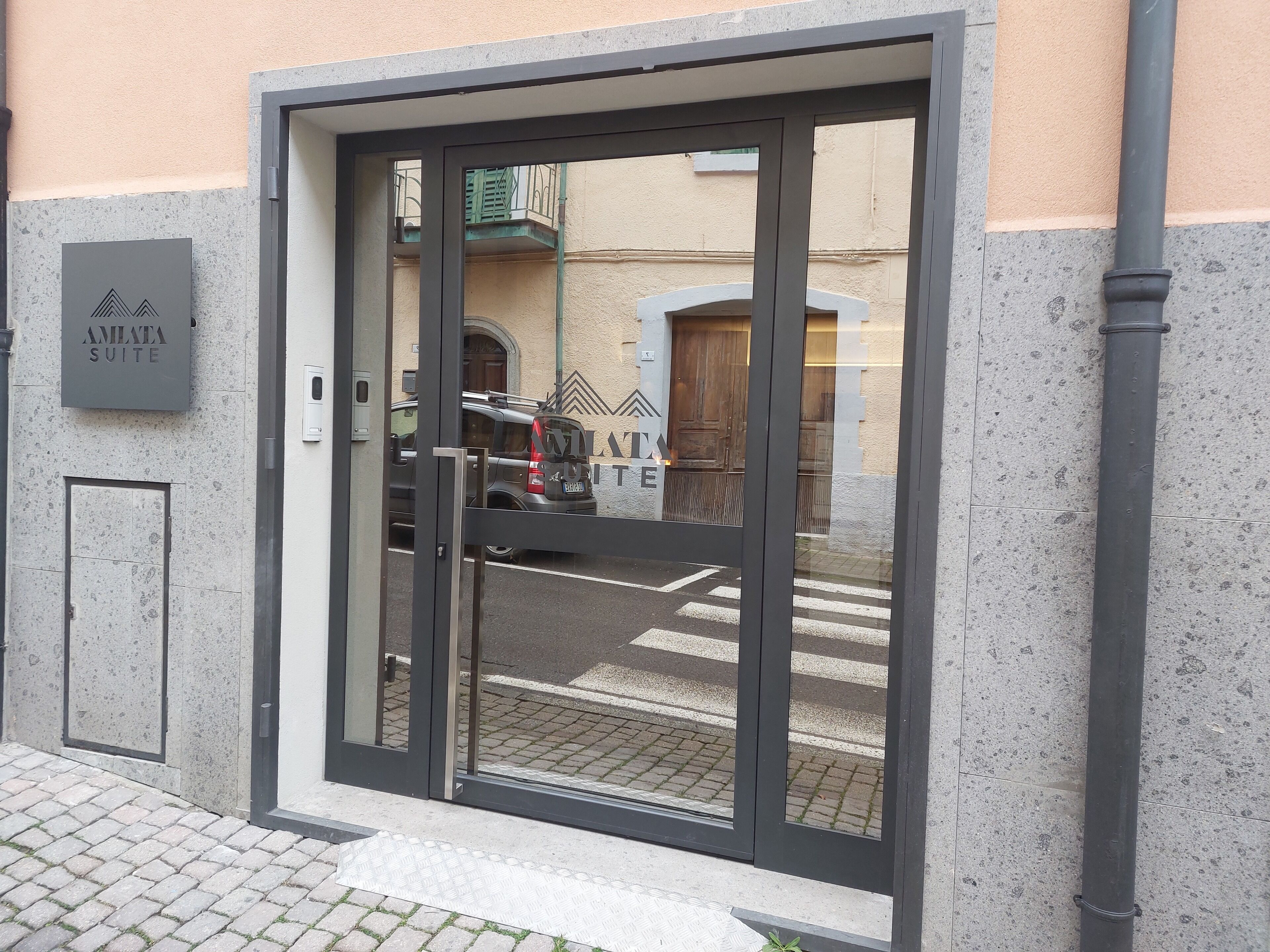 Ingresso moderno di un edificio con porta in vetro, un cartello con la scritta 'AMENA SUITE' e un veicolo parcheggiato all'esterno.