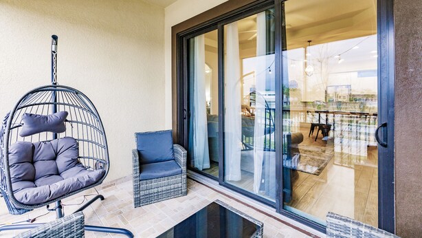 A balcony with a wicker chair and a sofa, a glass door leading to an indoor dining area.