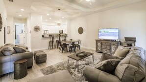 A spacious living room with a sofa, ottoman, coffee table, and a TV. There is a dining area with chairs and a table, and a kitchen in the background.