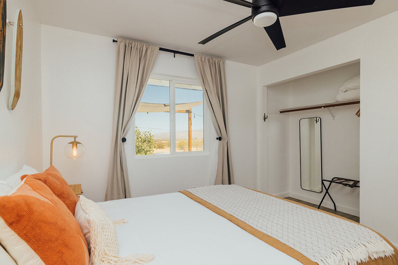Un dormitorio con cama, ventilador de techo, ventana con cortinas, espejo y una mesita de noche con lámpara.