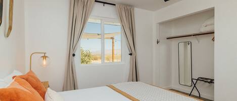 Un dormitorio con cama, ventilador de techo, ventana con cortinas, espejo y una mesita de noche con lámpara.
