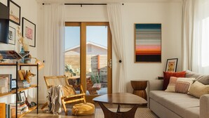 Un salon comprenant un canapé, un fauteuil, une petite table, une bibliothèque et une grande fenêtre avec des rideaux.