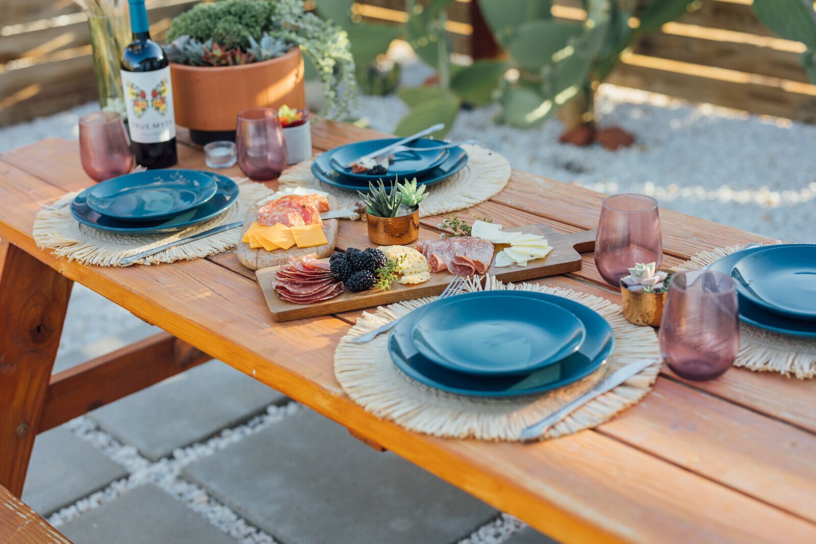 A wooden picnic table with blue plates, a wine bottle, and a variety of cheeses and meats.