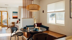 Un espace repas moderne avec une table ronde, des chaises, un luminaire suspendu et une grande fenêtre équipée de stores.