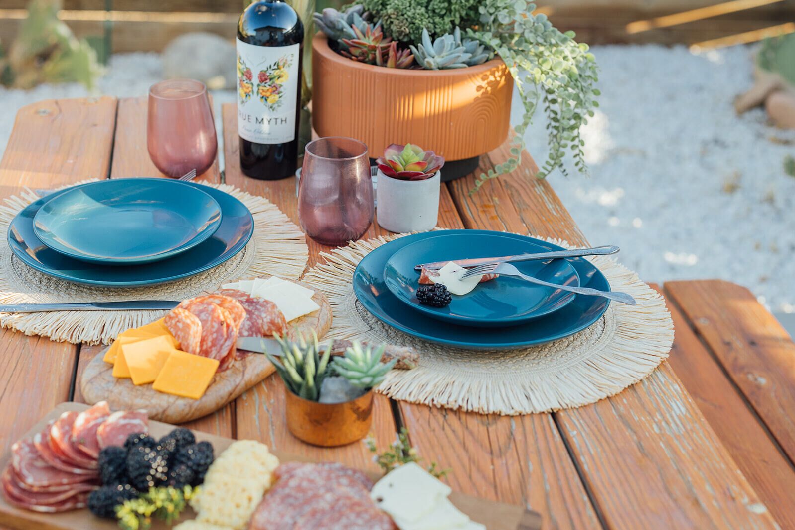 A wooden table with plates, cutlery, a bottle of wine, and a variety of cheeses and cured meats.