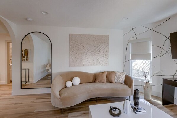 En moderne stue med en buet beige sofa, et stort spejl og et minimalistisk abstrakt billede.