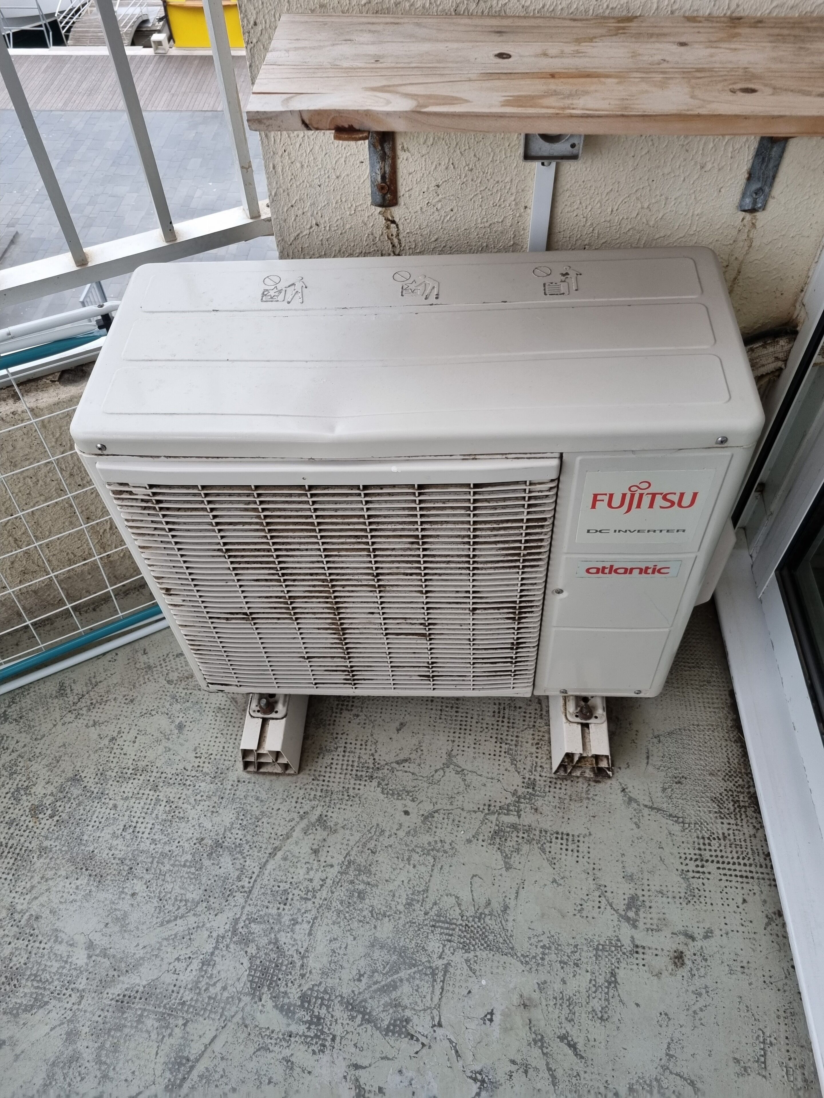 En hvid Fujitsu DC-inverter enhed monteret på en beton gulv, ved siden af et metal gelænder og en træplanke.