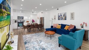 A modern living room with a blue sofa, a dining area with a wooden table, and a kitchen with white cabinets and a large painting on the wall. - Luminousness Reflection 4B2.5B Home (Torrance)