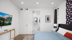 A modern bedroom with a bed, a TV, a desk, and a bathroom visible through an open door. - Luminousness Reflection 4B2.5B Home (Torrance)
