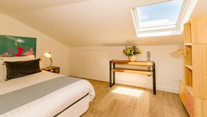 Ein Schlafzimmer mit Oberlicht, einem Bett mit grauer und weißer Bettdecke, einem Nachttisch mit Lampe, einem Holzbeistelltisch, einem an der Wand montierten Fernseher und einem Holzschrank.