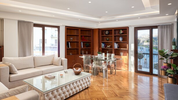 A spacious living room with a sofa, coffee table, and a glass-top dining table with chairs. There are wooden bookshelves and a large window with a view of the city. - Acropolis Prestige Residence (Athina)