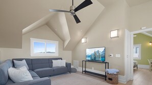 Un salón amplio con un sofá gris, un ventilador de techo, un televisor de pantalla plana y vistas a montañas a través de una ventana.
