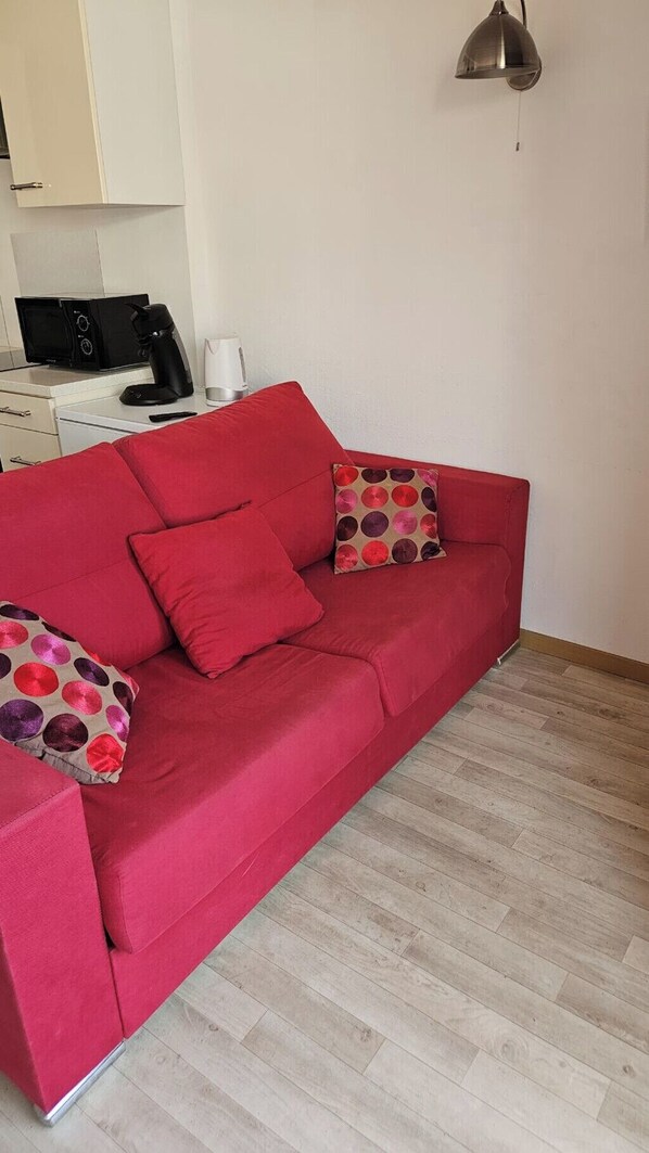 A red sofa with two patterned pillows, a coffee maker, and a microwave on a white cabinet in a room with wooden flooring.