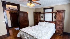 Une chambre avec un plancher en bois, un lit recouvert d’une couette blanche, un ventilateur de plafond et des meubles en bois.
