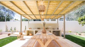 Un espacio cubierto para comer al aire libre con una mesa y bancos de madera, una lámpara colgante y una zona de cocina al fondo.