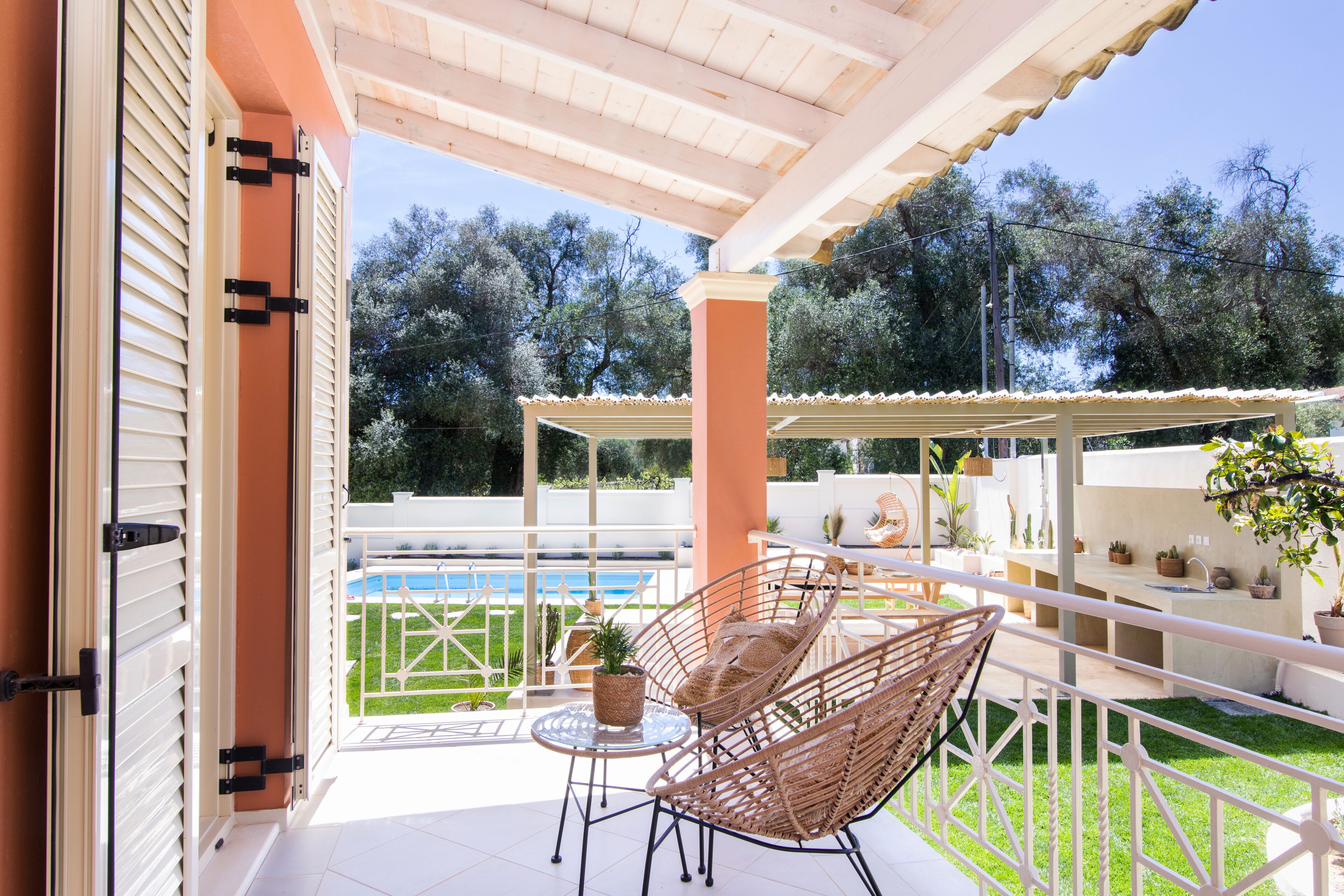 Une terrasse couverte aménagée avec des meubles en osier, une petite table ornée d’une plante, et une vue sur une piscine et la verdure environnante.