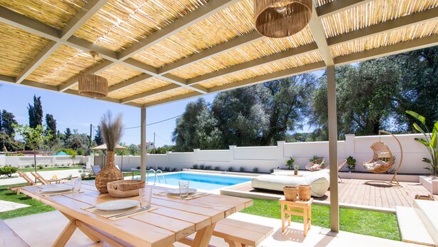 Un espacio al aire libre techado con piscina, muebles de madera y una zona de estar con cojines y una silla colgante.