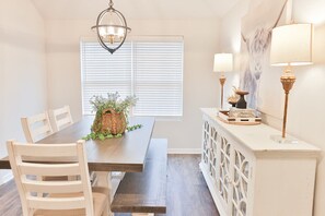 Een eetruimte met een houten tafel, witte stoelen, een hanglamp en een witte kast met decoratieve objecten.