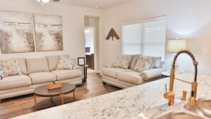 Ein modernes Wohnzimmer mit zwei beigefarbenen Sofas, einem Couchtisch und einer Küchenzeile mit Granitplatte und goldfarbenem Wasserhahn.