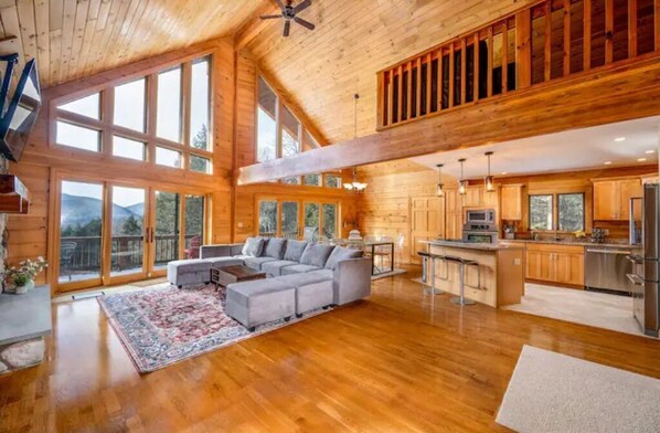 Relax in this open-plan living space with a large sofa, coffee table, and patterned rug, surrounded by large windows offering a view of the surrounding landscape.