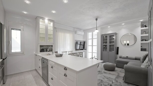 A modern kitchen with white cabinetry, a central island, and a dining area with a sofa and ottoman. - Belva Blue Glyfada Modern 2BR Walk to Ctr & Beach (Glifada)