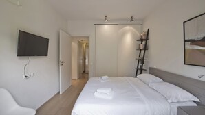 A modern hotel room with a large bed, a flat-screen TV mounted on the wall, a bookshelf, and a painting. - Sea Breeze Spacious Apt near Vouliagmeni Beach (Vouliagmeni)