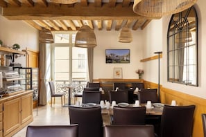 Un espace repas agrémenté de poutres en bois, de luminaires suspendus en osier et offrant une vue sur un bâtiment à travers une fenêtre.