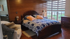 Quarto com cabeceira de madeira, colcha estampada, mesa de cabeceira com abajur e vista para um jardim pela janela.