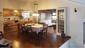 Uma cozinha espaçosa com armários de madeira, um espaço de refeições com mesa e cadeiras, e uma sala de estar com sofá e vista através de uma porta de vidro.