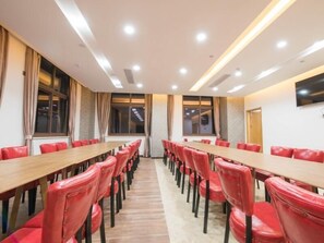 Un salón de conferencias con mesas largas y sillas rojas, ventanas amplias y un televisor de pantalla plana en la pared.